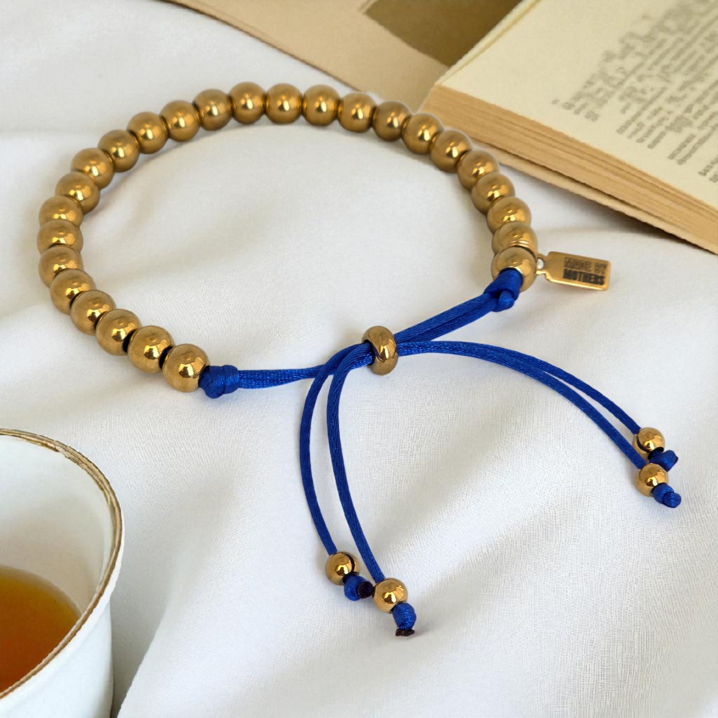 Bracelet with gold beads and blue cord on a white surface with a book and cup.