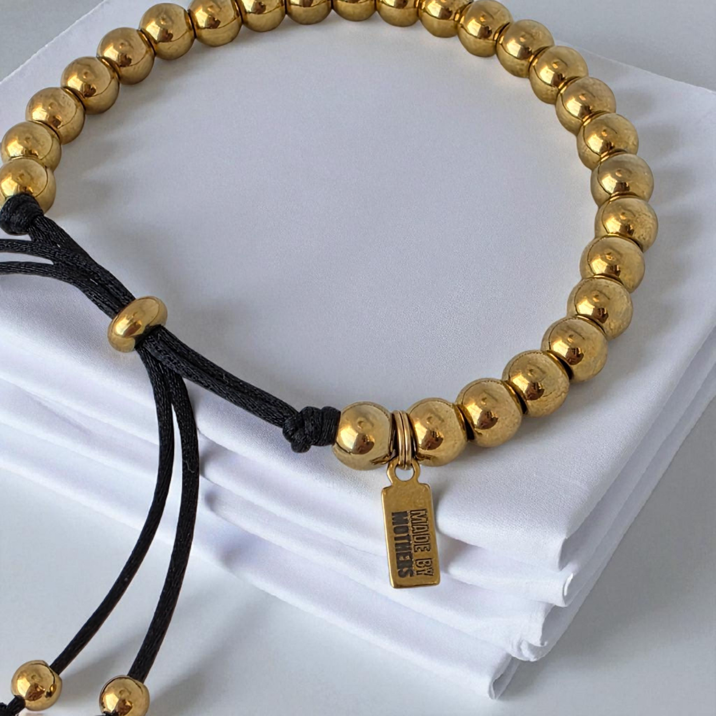 Gold beaded bracelet with a black cord on a white fabric background