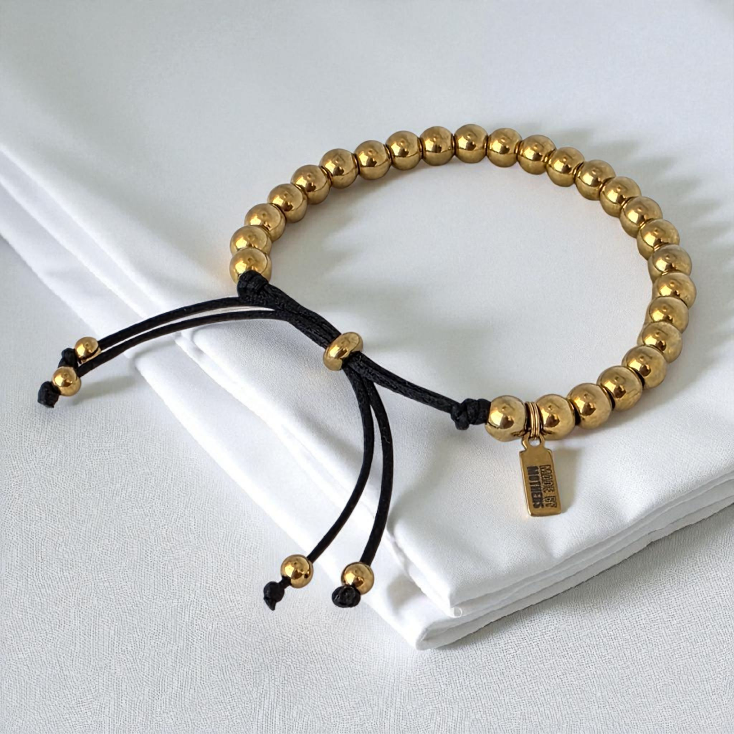 Gold beaded bracelet with a black cord on a white fabric background