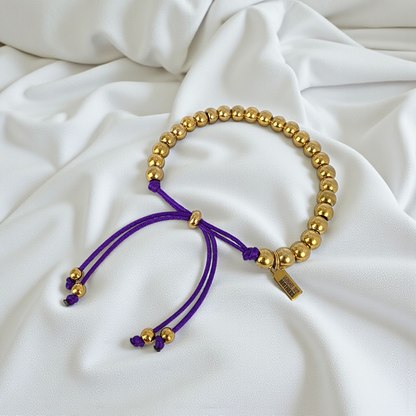 Gold beaded bracelet with purple cord on a white fabric background