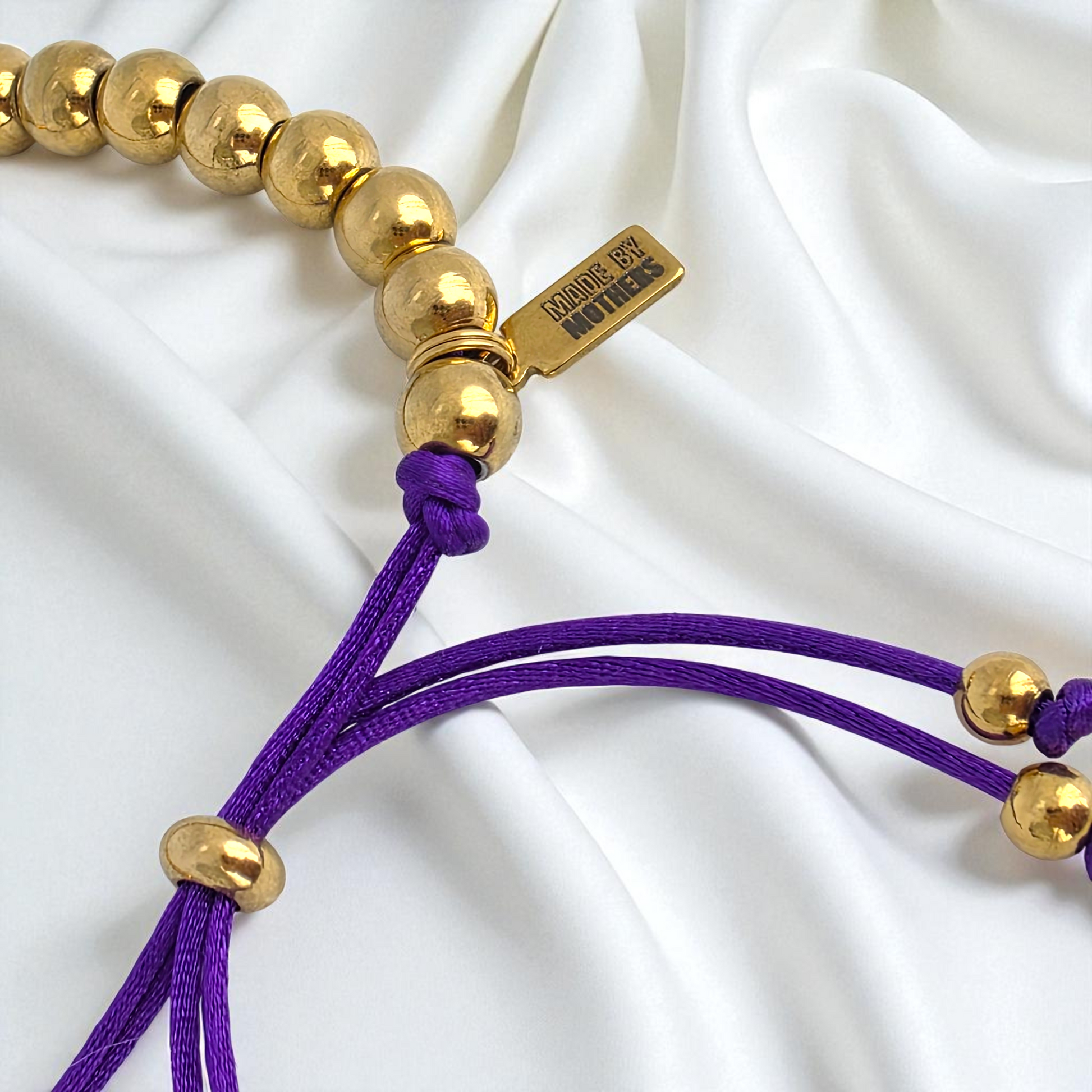 Gold beaded bracelet with purple cord on a white fabric background